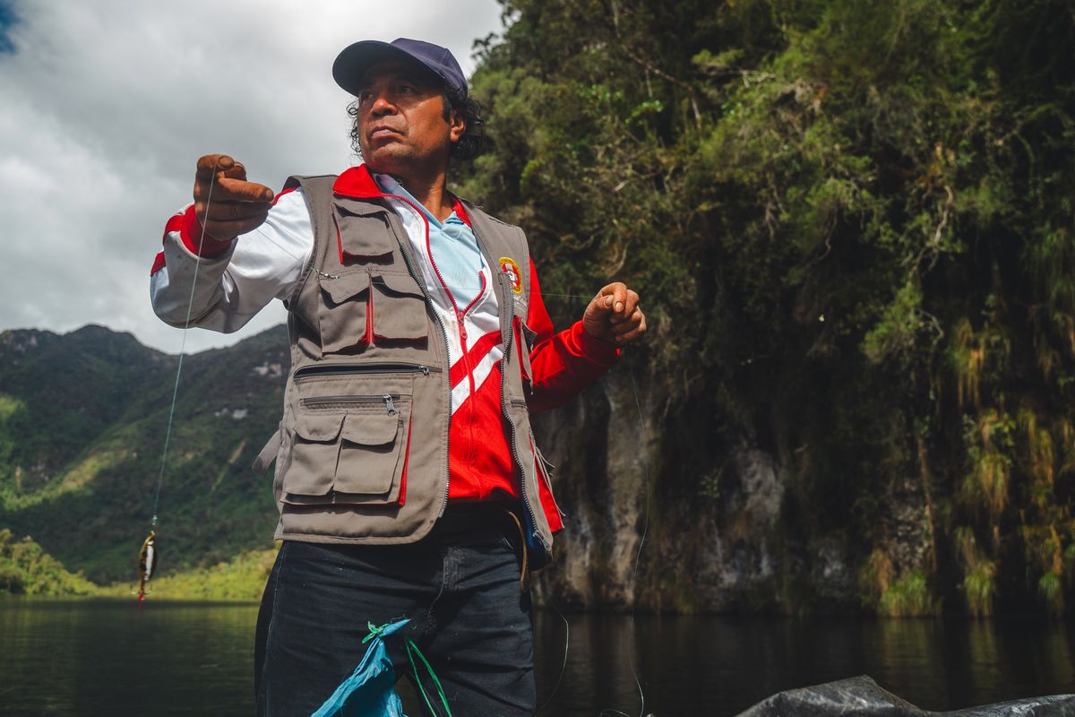 A local guide standing in a boat on Laguna de los Cóndores, handline fishing for trout with cliff walls and cloud forest behind him