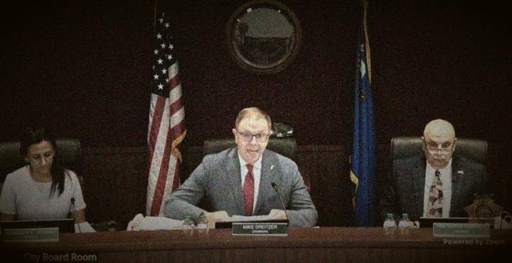 Grainy screenshot of three board members, including Chairman Mike Dreitzer, seated at a dais with flags behind them.