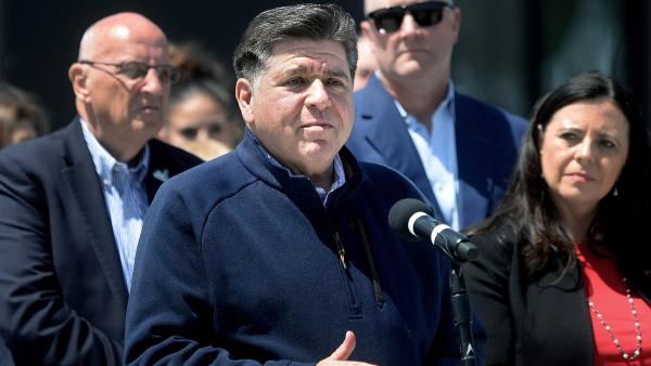 Illinois Governor JB Pritzker speaks at a microphone during a press conference with other officials standing behind him.