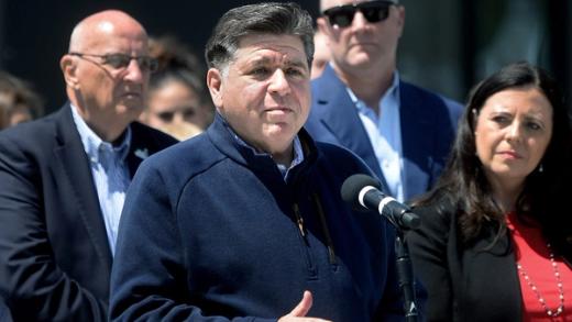 Illinois Governor JB Pritzker speaks at a microphone during a press conference with other officials standing behind him.