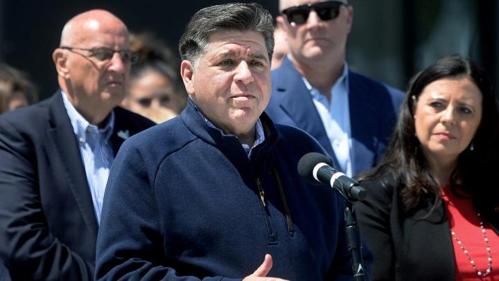 Illinois Governor JB Pritzker speaks at a microphone during a press conference with other officials standing behind him.