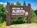Wooden roadside sign reading "Welcome to Alberta Wild Rose Country" with a pink rose illustration, set against green