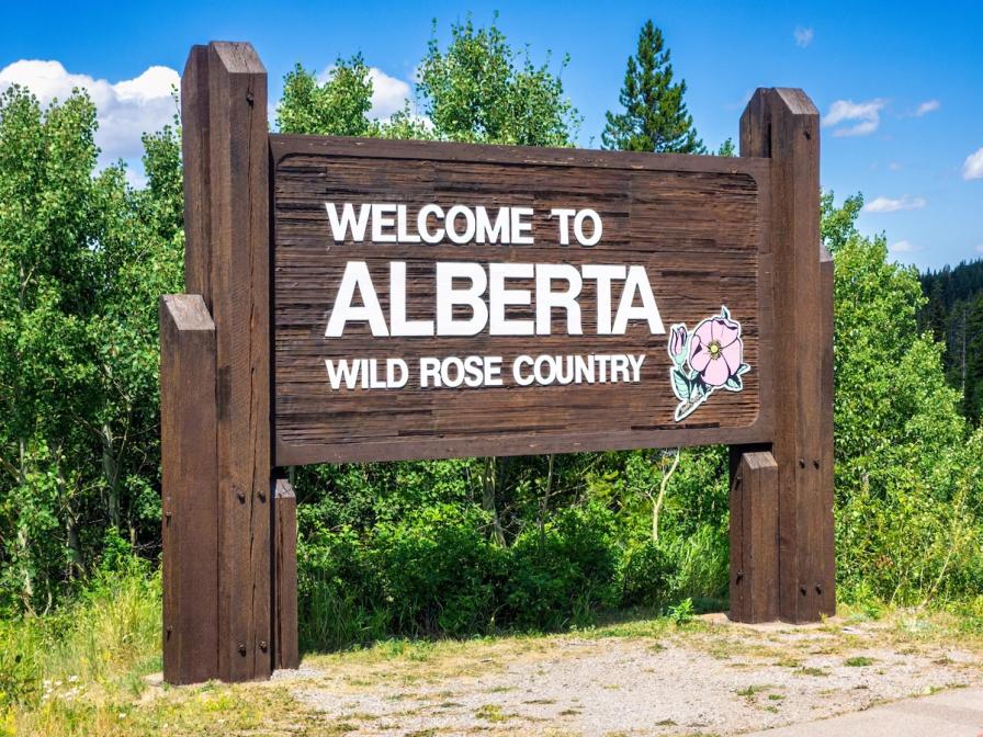Wooden roadside sign reading "Welcome to Alberta Wild Rose Country" with a pink rose illustration, set against green