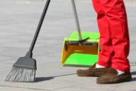 A person in red pants and brown loafers sweeps a paved ground with a black broom, with a green dustpan in the background.
