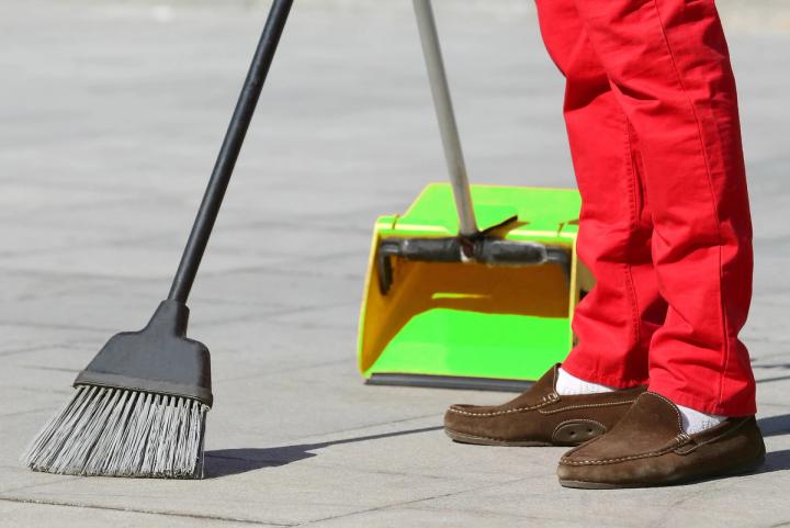 A person in red pants and brown loafers sweeps a paved ground with a black broom, with a green dustpan in the background.