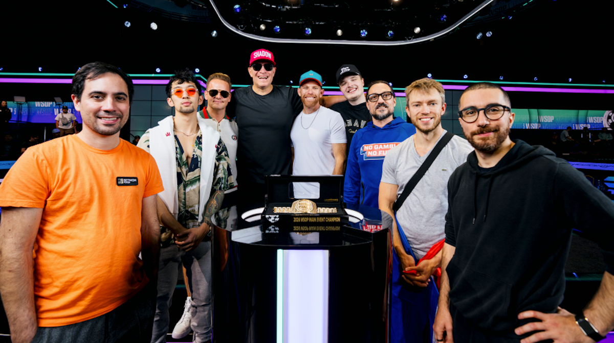 Nine men stand smiling together behind a podium displaying a gold championship bracelet at the WSOP Main Event.