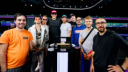 Nine men stand smiling together behind a podium displaying a gold championship bracelet at the WSOP Main Event.