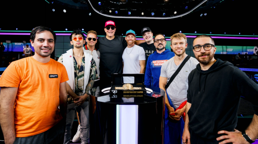 Nine men stand smiling together behind a podium displaying a gold championship bracelet at the WSOP Main Event.
