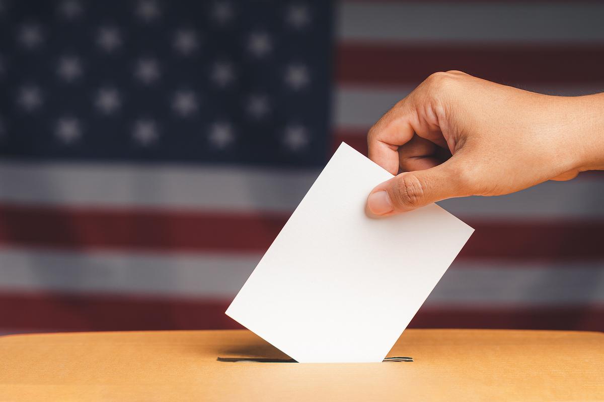 A hand casting a ballot into a ballot box against a blurred US flag background.
