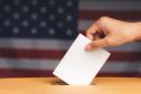 A hand casting a ballot into a ballot box against a blurred US flag background.