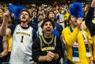 Excited Michigan sports fans cheering and shouting in a crowded stadium, waving blue pom-poms.