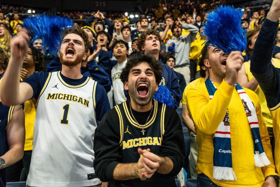 Excited Michigan sports fans cheering and shouting in a crowded stadium, waving blue pom-poms.