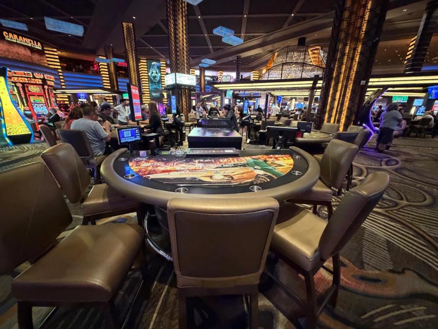 An empty card table with leather chairs on a busy casino floor with slot machines and people in the background.
