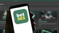 A hand holding a smartphone displaying the bet365 app logo, with a blurred sports betting website in the background.