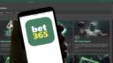 A hand holding a smartphone displaying the bet365 app logo, with a blurred sports betting website in the background.