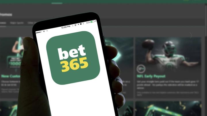A hand holding a smartphone displaying the bet365 app logo, with a blurred sports betting website in the background.