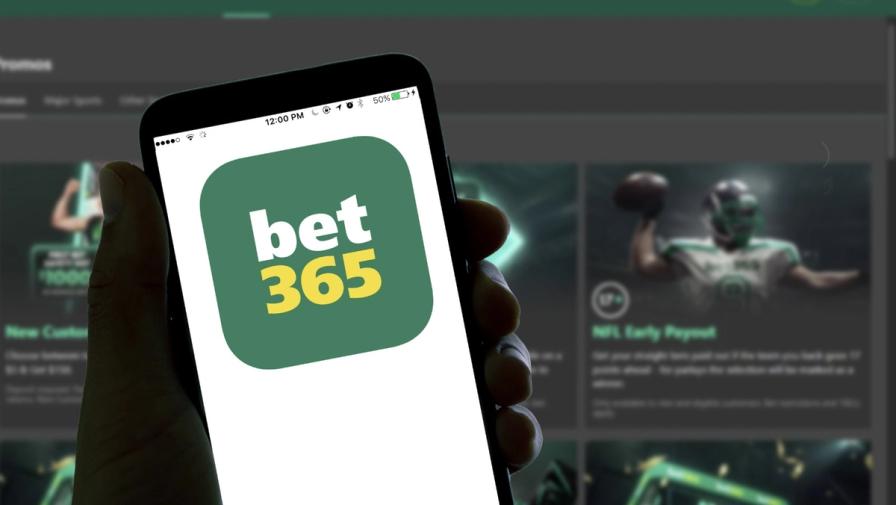 A hand holding a smartphone displaying the bet365 app logo, with a blurred sports betting website in the background.