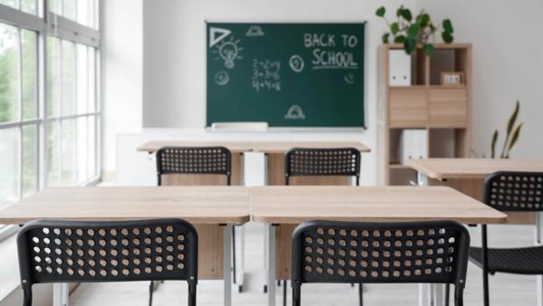 An empty classroom with wooden desks, black chairs, and a green chalkboard in the background reading Back to