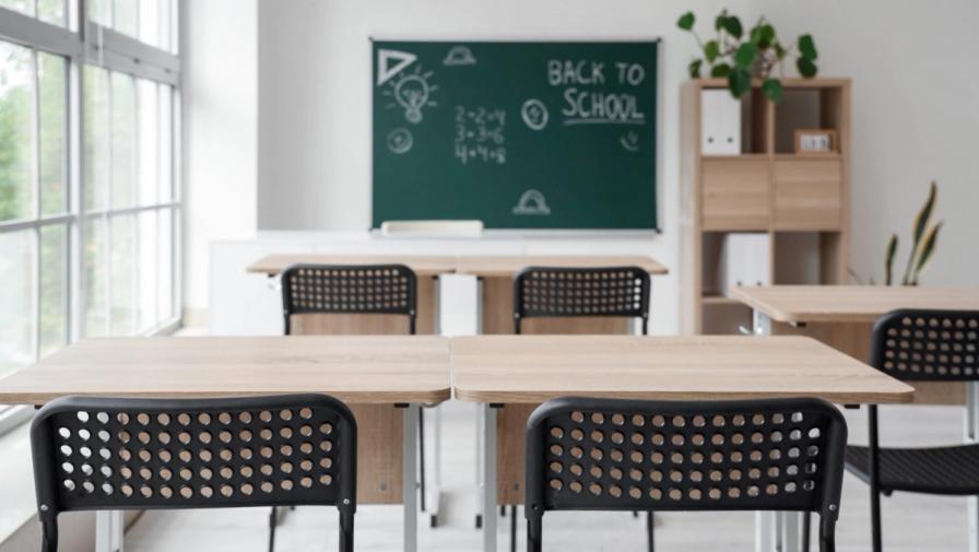 An empty classroom with wooden desks, black chairs, and a green chalkboard in the background reading Back to