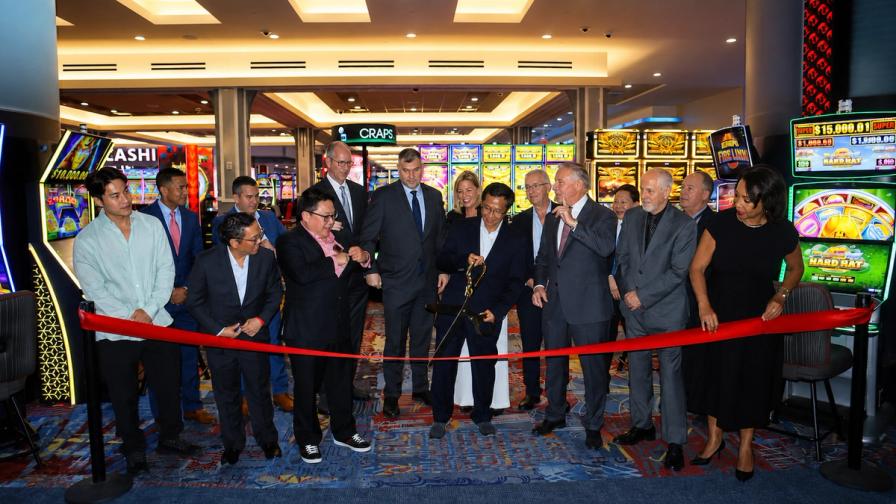 A group of people participate in a ribbon-cutting ceremony on a casino floor surrounded by slot machines.