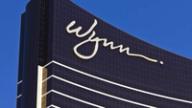 The white cursive Wynn logo on the dark glass facade of a Wynn resort building against a clear blue sky.