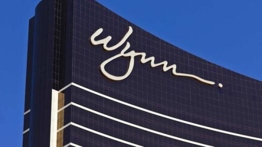 The white cursive Wynn logo on the dark glass facade of a Wynn resort building against a clear blue sky.