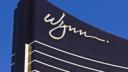 The white cursive Wynn logo on the dark glass facade of a Wynn resort building against a clear blue sky.