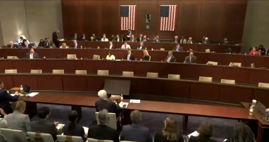 A US House committee hearing in progress, with members seated at tiered desks below two American flags.