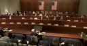 A US House committee hearing in progress, with members seated at tiered desks below two American flags.