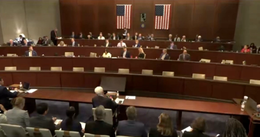 A US House committee hearing in progress, with members seated at tiered desks below two American flags.