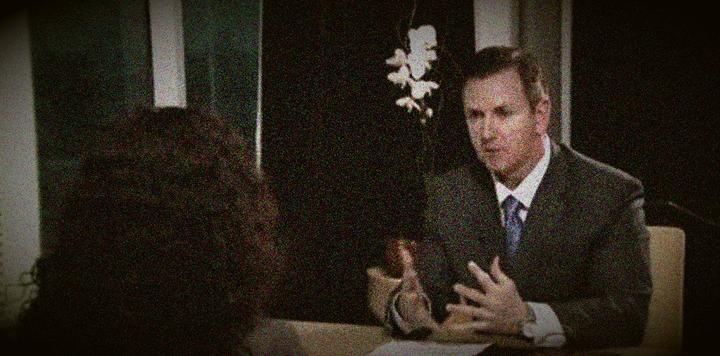 Grainy image of a man in a suit gesturing while speaking to someone with dark curly hair across a table.