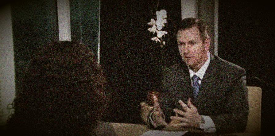 Grainy image of a man in a suit gesturing while speaking to someone with dark curly hair across a table.