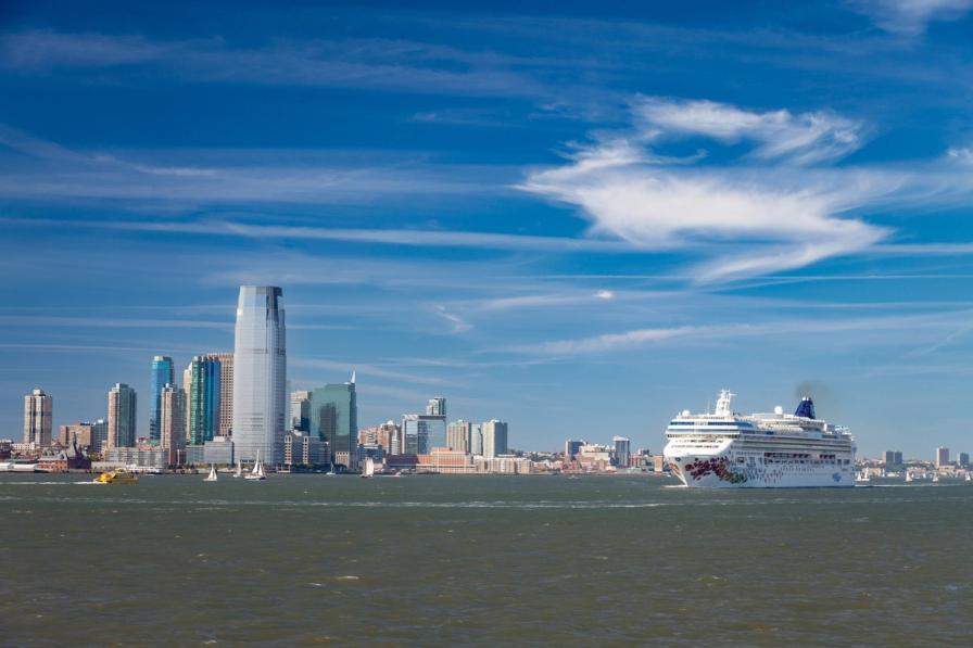A cruise ship sails past the Jersey City skyline on a sunny day.