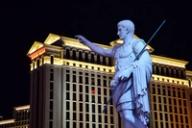 A statue of Julius Caesar pointing forward, with the illuminated Caesars Palace hotel in Las Vegas in the background at night.