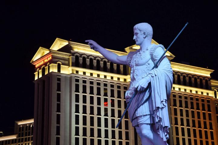 A statue of Julius Caesar pointing forward, with the illuminated Caesars Palace hotel in Las Vegas in the background at night.