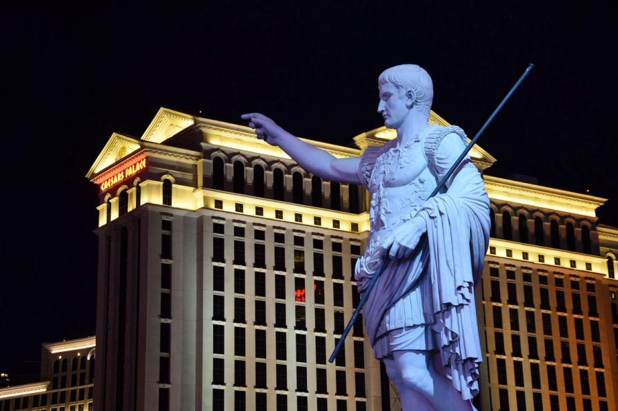 A statue of Julius Caesar pointing forward, with the illuminated Caesars Palace hotel in Las Vegas in the background at night.