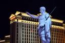 A statue of Julius Caesar pointing forward, with the illuminated Caesars Palace hotel in Las Vegas in the background at night.