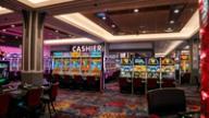 Brightly lit casino floor featuring rows of colorful slot machines with red chairs and a cashier sign in the background.