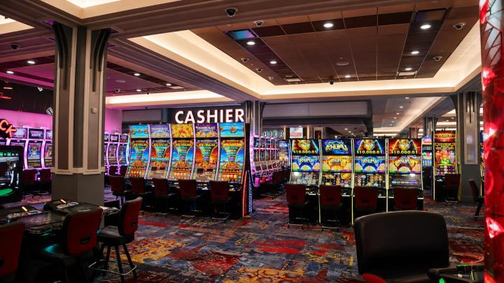 Brightly lit casino floor featuring rows of colorful slot machines with red chairs and a cashier sign in the background.