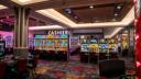 Brightly lit casino floor featuring rows of colorful slot machines with red chairs and a cashier sign in the background.