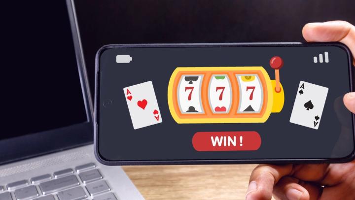 A hand holding a smartphone displaying an online slot machine game with triple sevens and a WIN button next to a laptop.