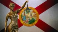A bronze statue of Lady Justice holding scales in front of the Florida state flag.