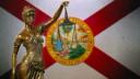 A bronze statue of Lady Justice holding scales in front of the Florida state flag.