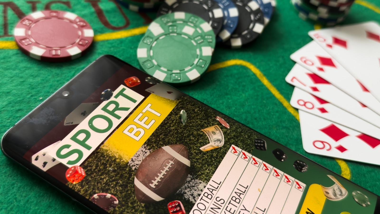 A smartphone displaying a sports betting app on a green casino table next to poker chips and playing cards.