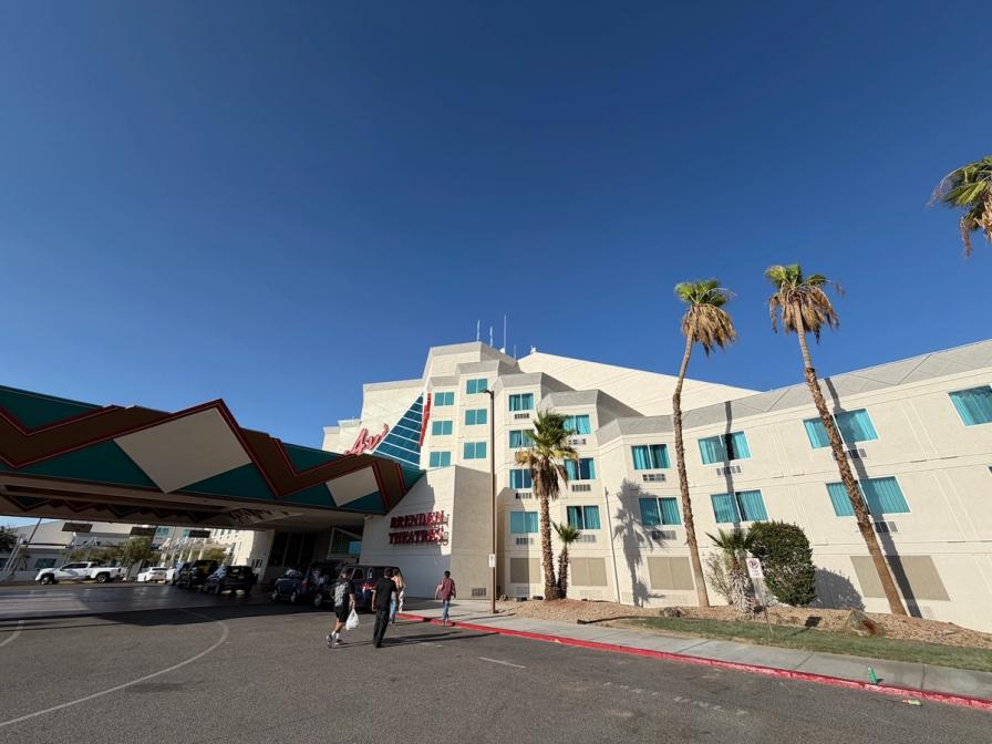 Exterior of a white hotel and casino building with palm trees, a colorful canopy, and people walking toward the entrance.