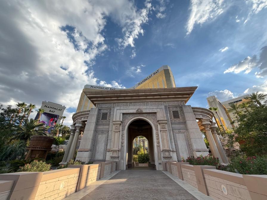 An ornate stone archway and bridge leading toward the golden Mandalay Bay hotel in Las Vegas under a cloudy blue sky.