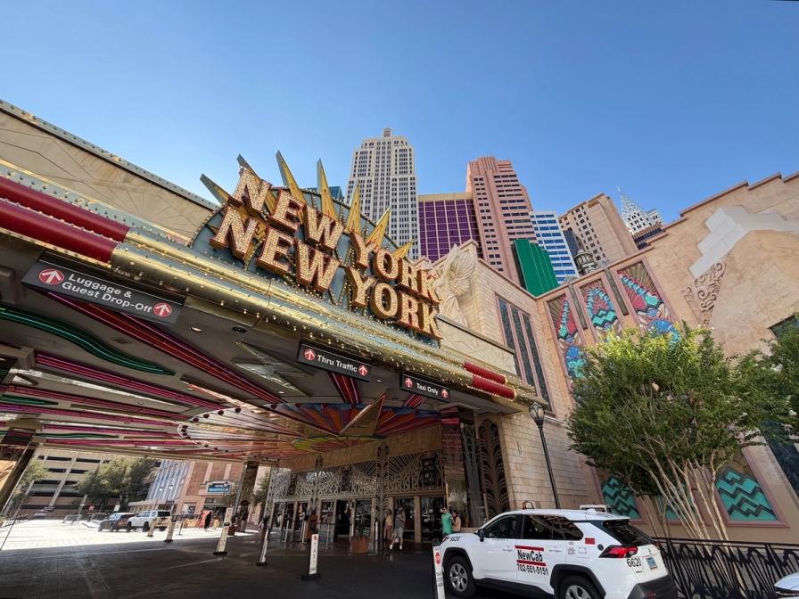Entrance of the New York-New York Hotel & Casino in Las Vegas, showing the marquee sign, replica skyline, and a white taxi.