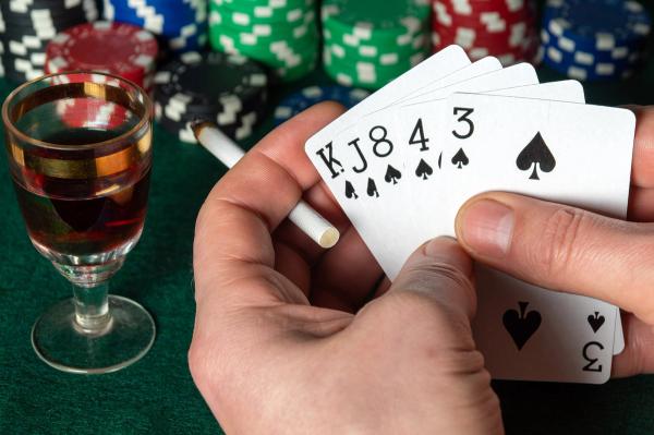Hands holding a spade poker hand and a cigarette, next to a glass of alcohol and poker chips on a green felt table.