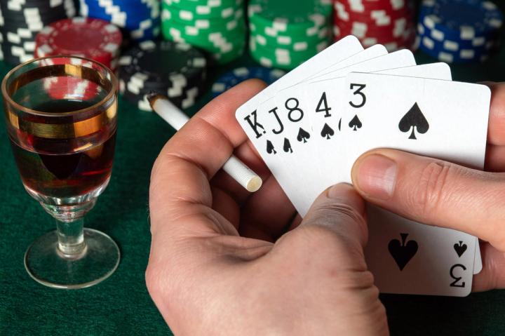 Hands holding a spade poker hand and a cigarette, next to a glass of alcohol and poker chips on a green felt table.