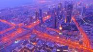 Aerial view of Dubai's skyline and glowing highway interchanges at twilight, bathed in purple and orange light.